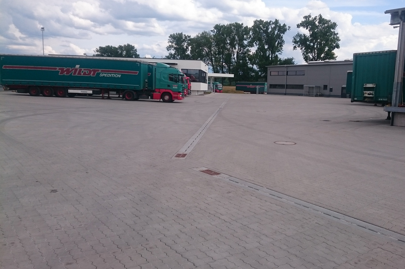 Grüne und rote LKWs stehen auf gepflasterter Logistikfläche. Pfuhler Rinne ist verlegt.