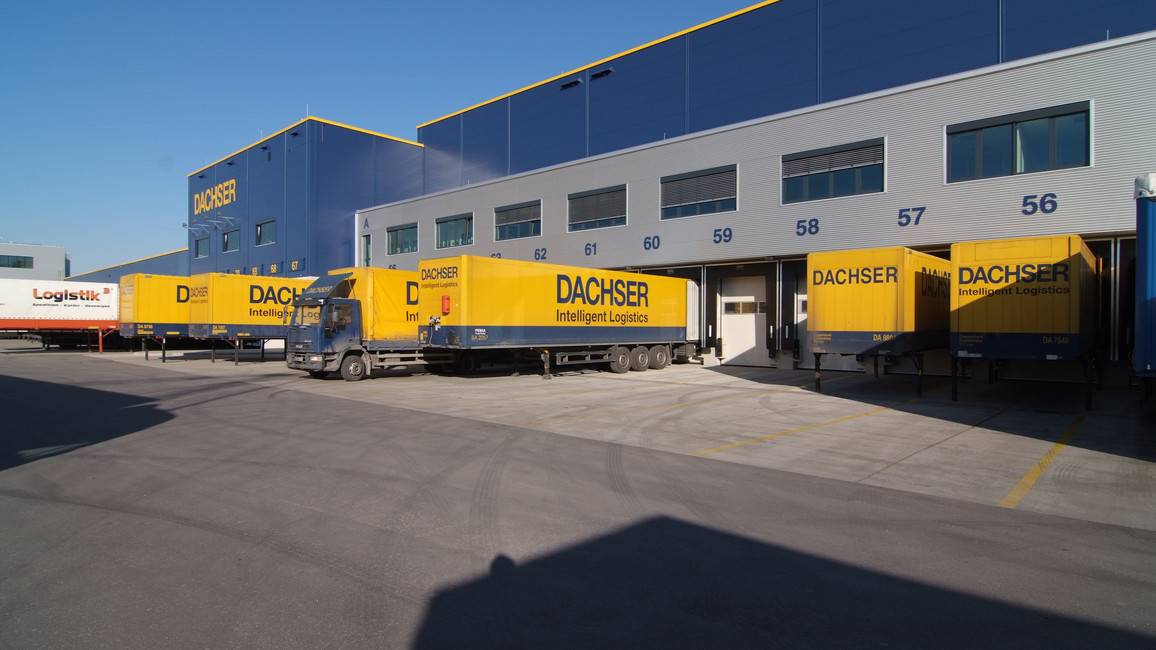 Mehrere gelbe Dachser LKWs stehen auf einer Logistikfläche und werden beladen. Die Sonne scheint und der Himmel ist blau.