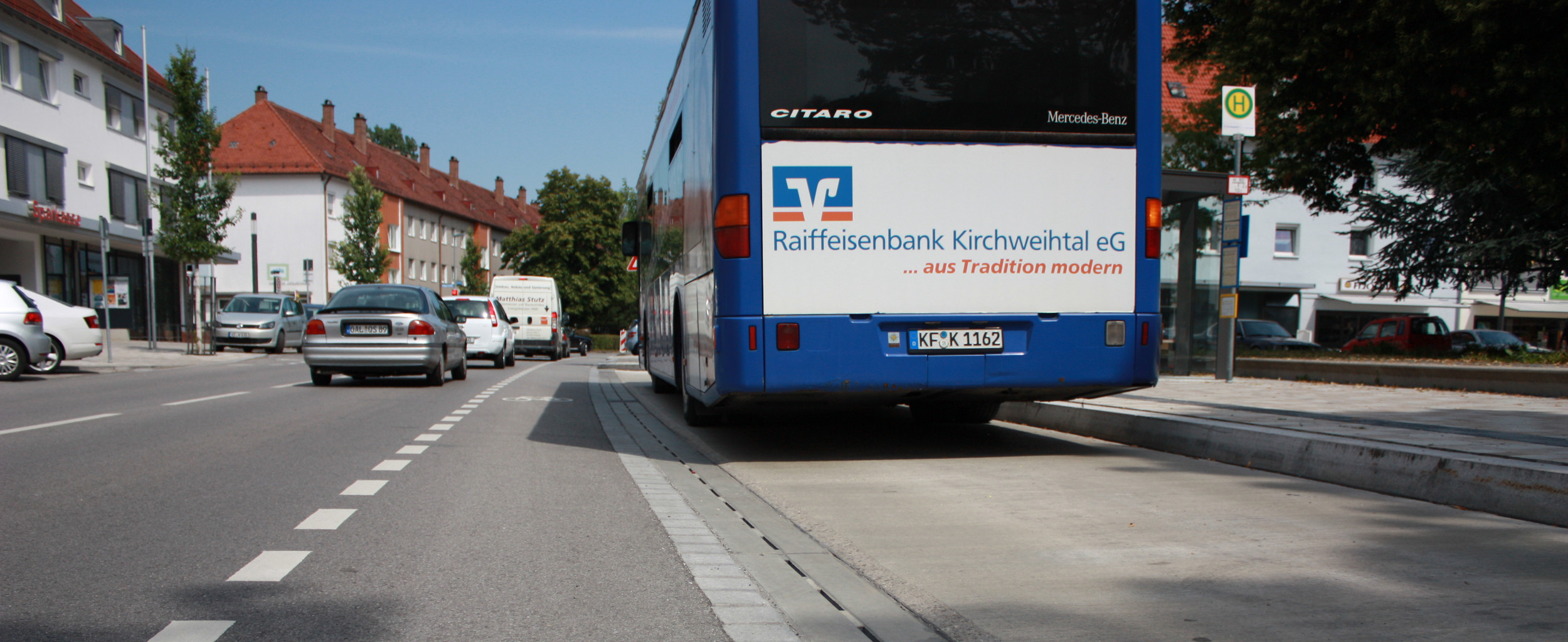 Hauptstraße in der Stadt. Rechts steht ein blauer Bus an einer Haltestelle. Eine City Drain Rinne legt zwischen der Fahrbahn und der Haltestelle.
