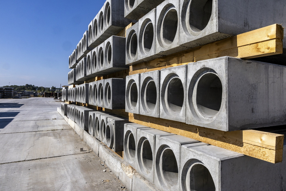 Betonrinnen stehen palettenweise übereinandergestapelt auf einem Betriebsgelände. Die Sonne scheint, der Himmel ist blau.