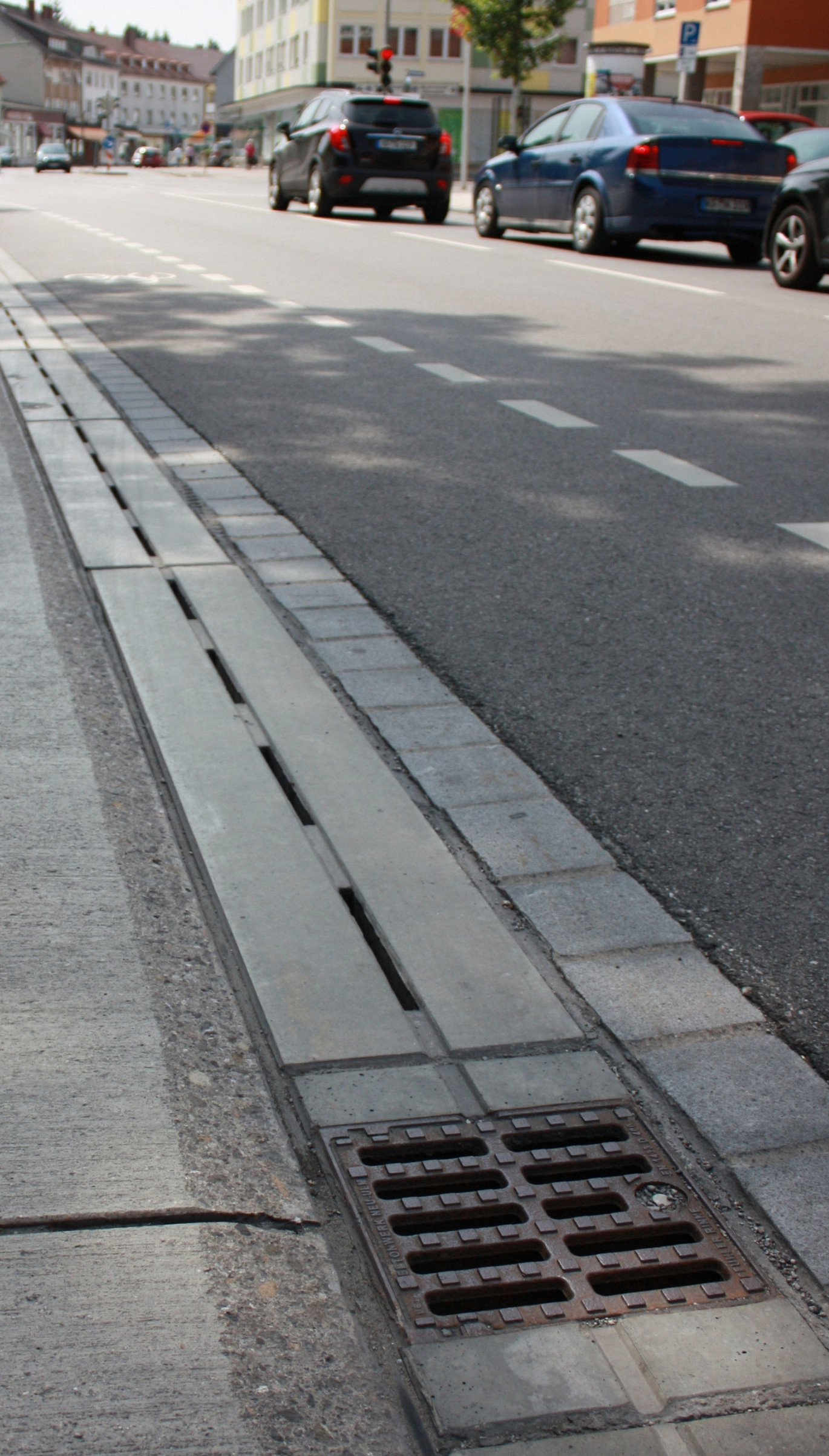 Hauptstraße in der Stadt. Autos stehen vor einer roten Ampel. Die Sonne scheint und eine Baum wirft einen großen Schatten auf die Straße und die darin verbaute Entwässerungsrinne.