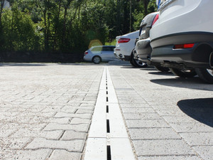 City Drain Rinne auf Parkplatz verlegt. Parkende Autos stehen vor der Rinne.