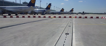 Flugplatz, auf dem einige Lufthansa Flugzeuge stehen. Eine Pfuhler Rinne  im Vordergrund vor einer weiß-roten Markierung. Die Sonne scheint.