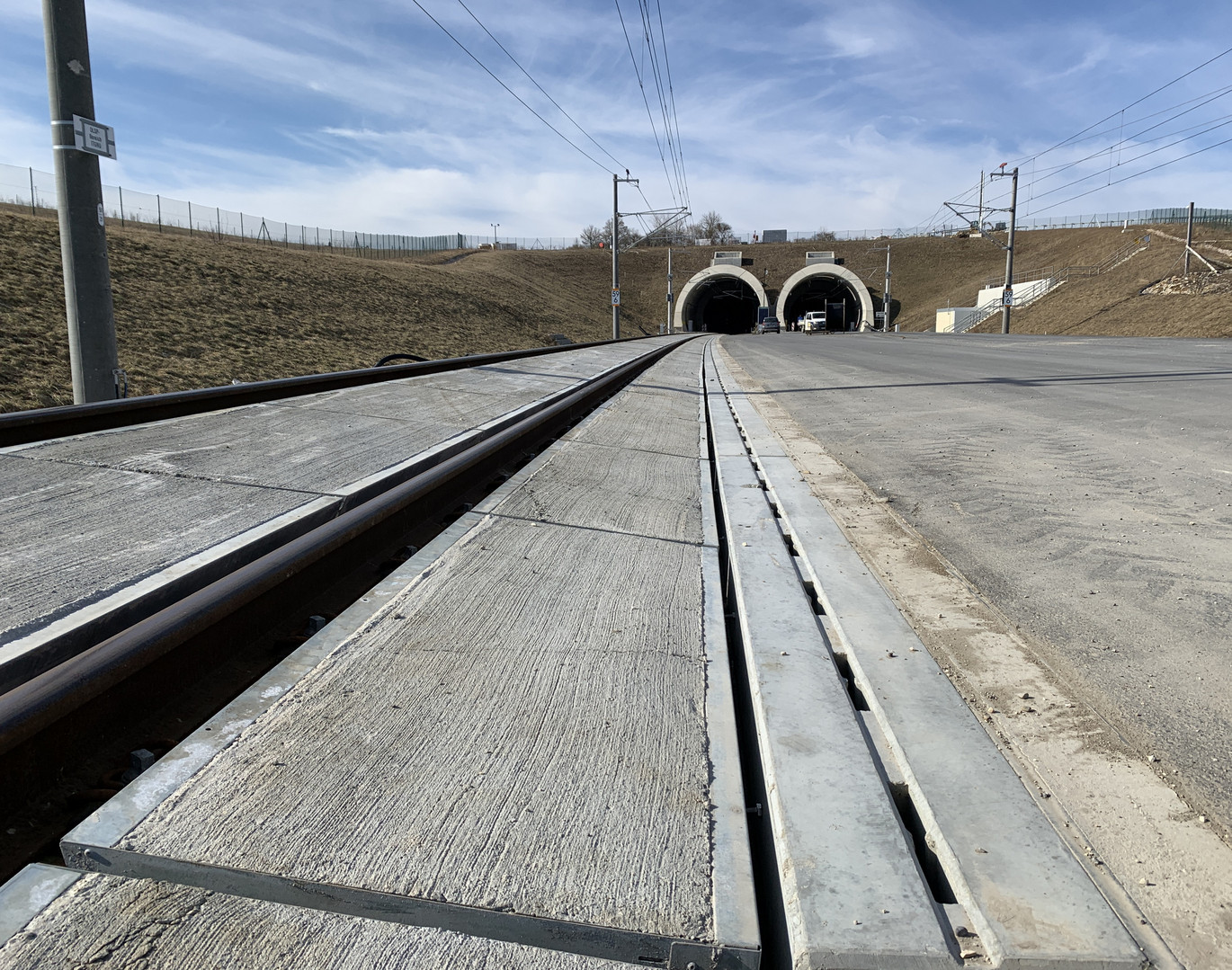 Zwei Tunnelöffnungen, links für Schienenverkehr und rechts für PKWs, durch die Mitte zieht sich eine Betonrinne. Der Himmel ist blau.