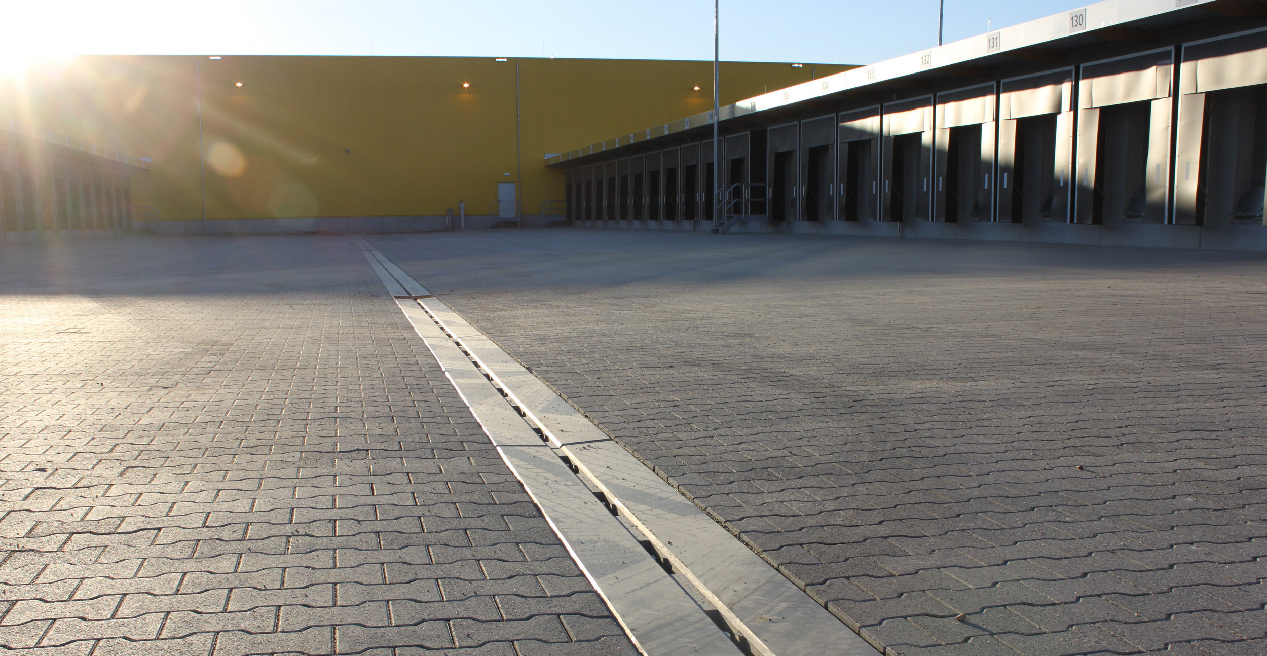 Betonrinne zieht sich über gepflasterte Logistikfläche. Gelbe Wand im Hintergrund. Die Sonne scheint.