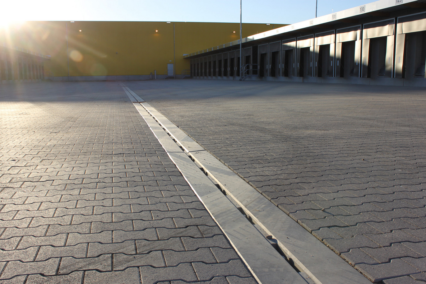 Betonrinne zieht sich über gepflasterte Logistikfläche. Gelbe Wand im Hintergrund. Die Sonne scheint.