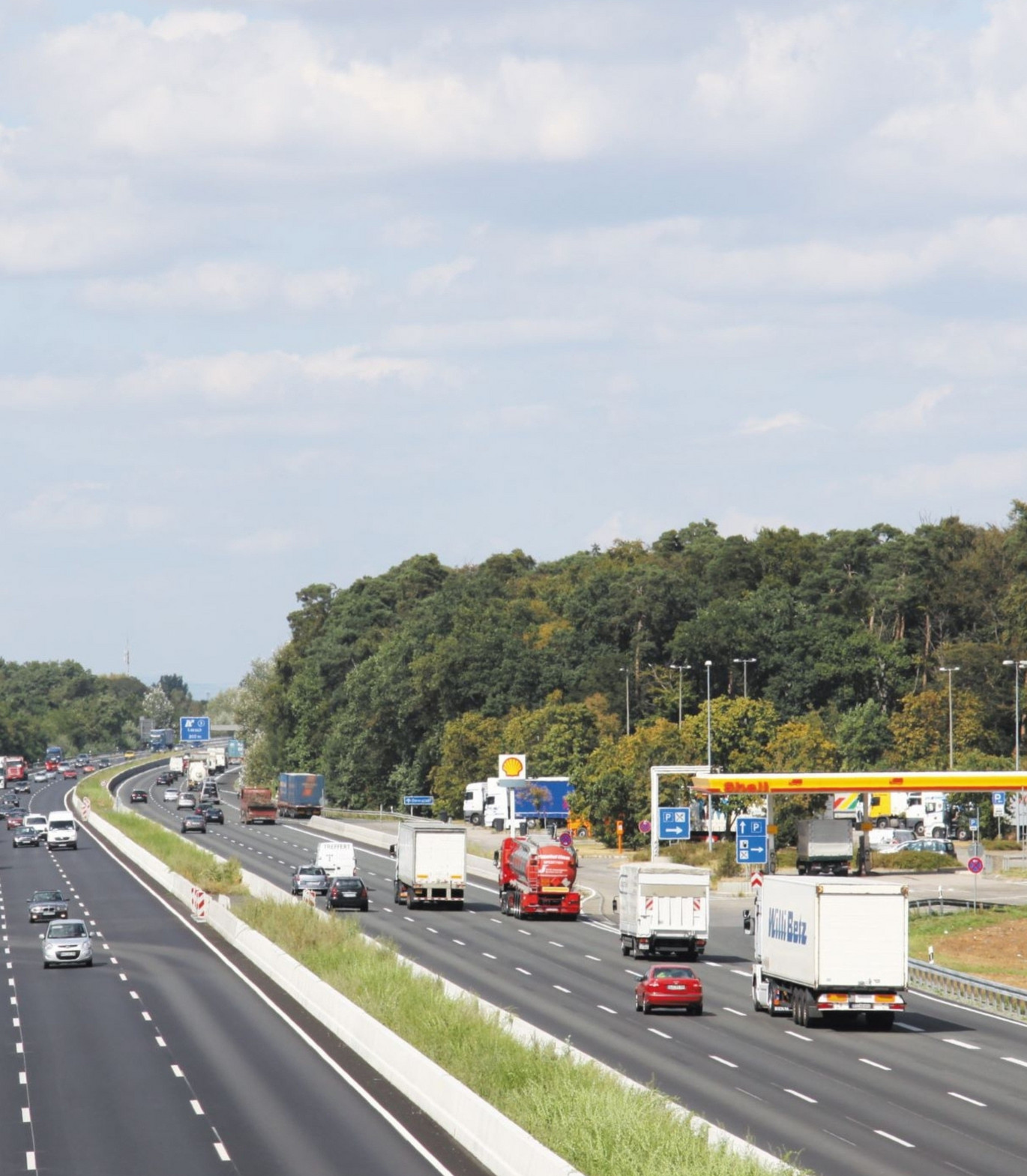 dreispurige Autobahn aus der Vogelperspektive. Rechts eine Shell-Tankstelle. Rund herum grüne Bäume und blauer Himmel.