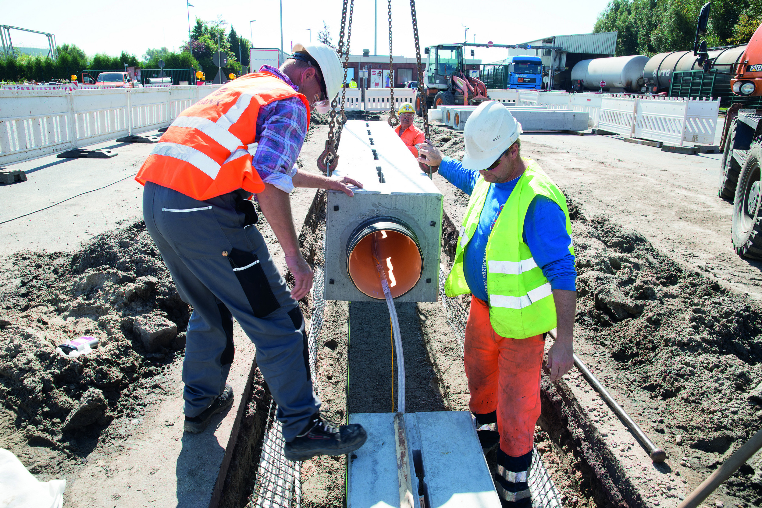 Drei Männer in Sicherheitskleidung verlegen eine CHEM 300 Rinne auf einer Baustelle. Im Hintergrund Baustellenfahrzeuge, LKWs und Absperrungen.